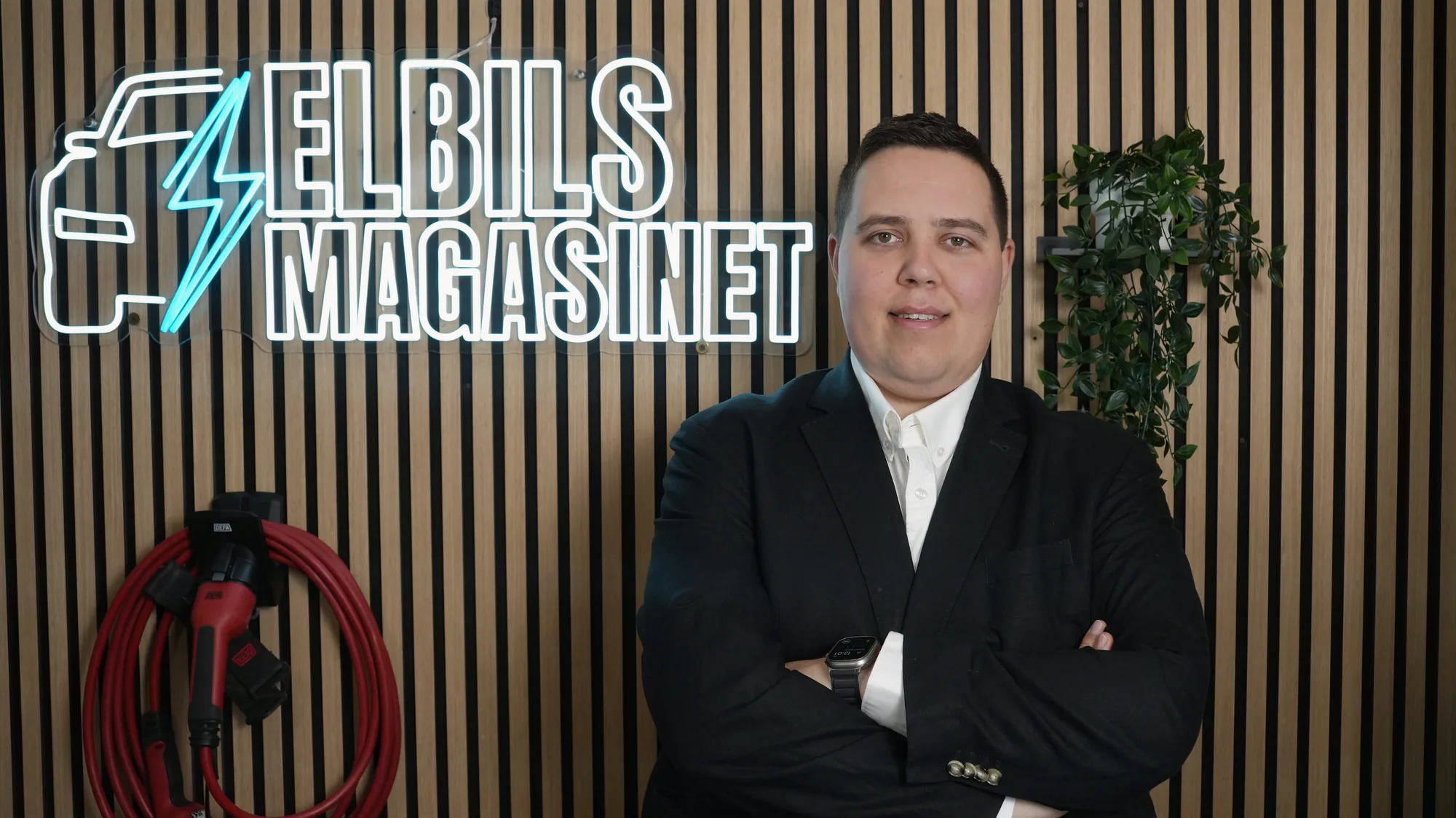 Christoffer Gullin i mörk kavaj med armarna i kors framför Elbilsmagasinets neonskylt i studion
